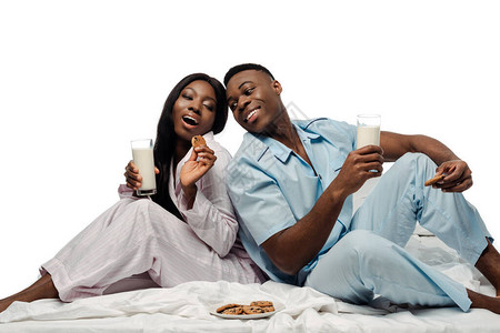 快乐的非洲夫妇吃饼干和牛奶在床上用睡衣孤立图片