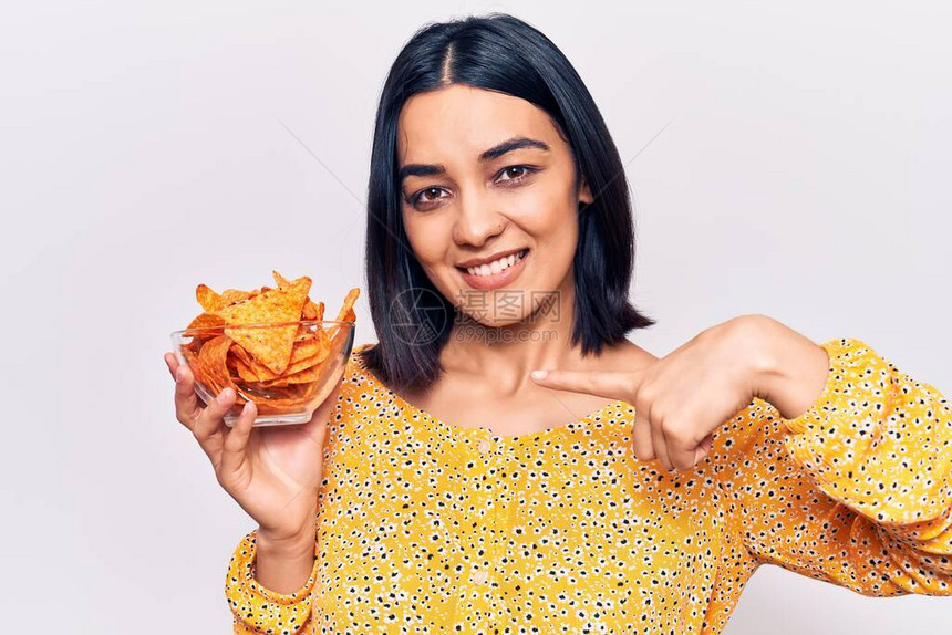 年轻美丽的拉丁女人拿着玉米薯片指向一个笑着快乐和骄傲的图片