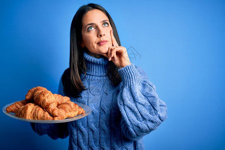 蓝眼睛的年轻女子在孤立的背景下拿着甜羊角面包吃早餐图片