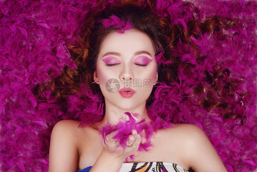 一个有着专业紫色妆容的漂亮女孩对着镜头送上一个飞吻该模型位于紫色羽毛中粉红色背景上的时尚照片卷图片