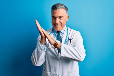中年英俊的灰发医生穿着外套和蓝色听诊器鼓掌快乐背景图片