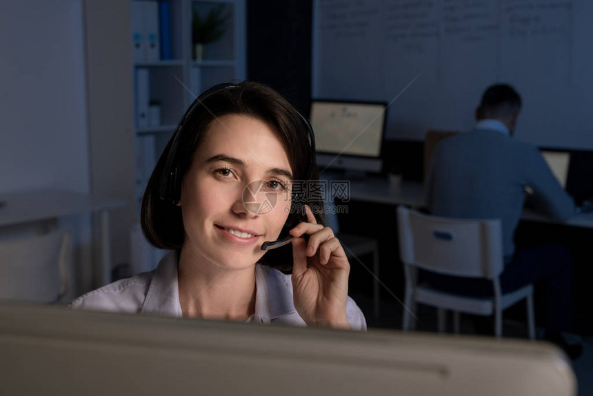 漂亮的年轻笑的接线员戴耳头巾看着你同时在办公室图片
