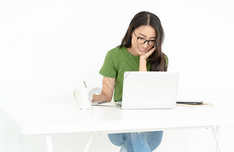 快乐的年轻亚洲少女戴着眼镜在家中使用笔记本电脑时在笔记本上记笔图片