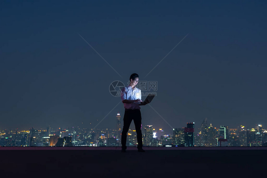 亚洲商人站在大楼的顶部使用笔记本与城市夜景商业成图片