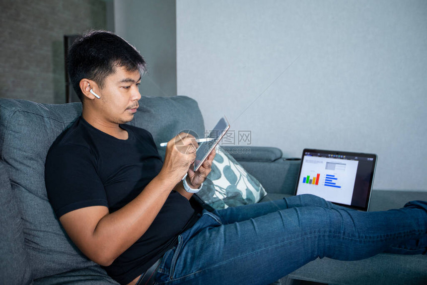 亚洲商人使用无线耳机笔记本和平板在家工作他为在家中举行的网上会议准备图表和信息校图片