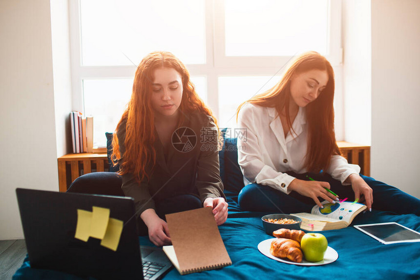 两名红发学生在家学习或准备考试年轻女在靠近窗户的宿舍床上做作业有笔记本食品书籍平板电脑笔记图片