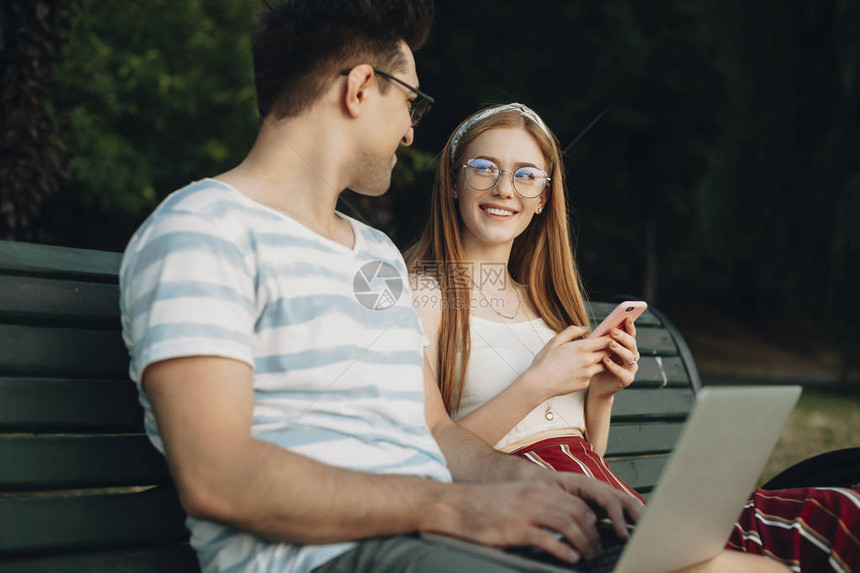 年轻的红头发女孩看着她的男朋友使用智能手机的雀斑两个年轻学图片