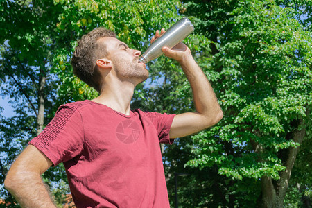 人在树叶背景中喝水口渴的家伙在公园户外锻炼后喝水人从多个金属瓶热阳光图片