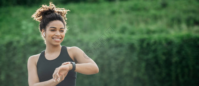 在户外运动带着无线耳机微笑的非洲女孩在绿公园背景上检查健身手镯关闭空图片