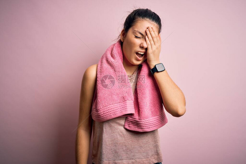 身穿运动服和毛巾穿着粉红色背景的年轻漂亮的棕发女运动员疲惫地用手遮住半张图片