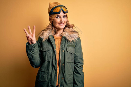 身穿雪运动服和滑雪护目镜的中年金发美女滑雪女郎展示并举起三指图片