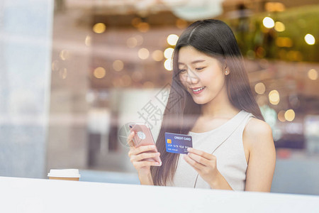年轻的亚洲女使用带手机的信用卡在咖啡店或窗镜旁的合作空间进行网上购物技术货币钱包和在线支付概念背景图片
