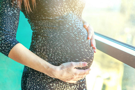 关闭怀孕的腹部孕妇站在家窗边的蓝墙附近产妇怀孕柔情父母身图片
