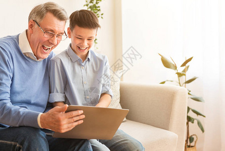 喜悦的祖父和男孩使用笔记本电脑观看喜剧电影一起坐在家里的库奇口背景图片