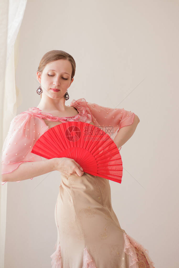 一个美丽的年轻女子在米色背景上的米色和粉红色连衣裙的肖像她正在用一把红扇子跳弗拉门戈图片