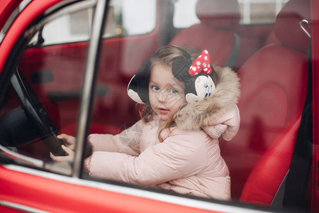 库存照片的可爱的小女孩在米奇耳罩和冬衣坐在红色汽车的司机座位上她在降雪下透过图片
