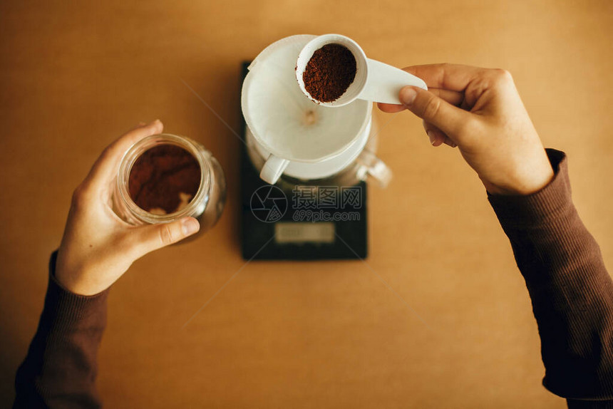 手倒在过滤器顶部视图中的磨碎的咖啡准备替代咖啡冲泡v60玻璃水壶背景上拿着勺子和磨咖啡的图片