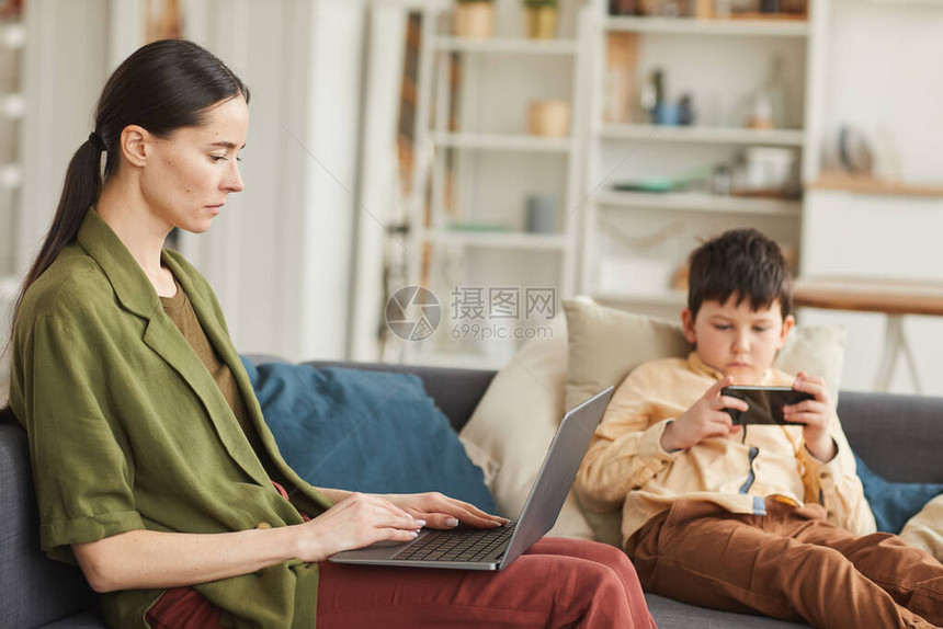 现代成年妇女在家里工作时用笔记本电脑与儿子一起在背景复制空间上玩移动游图片