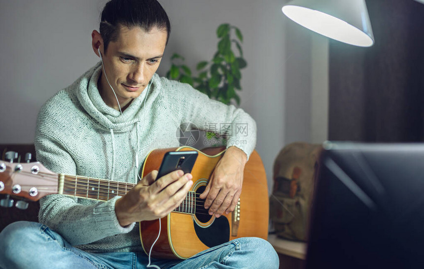 一位年轻的音乐家正在使用手机应用程序在线课程中学习弹奏原声吉他一个男人正在家里的图片