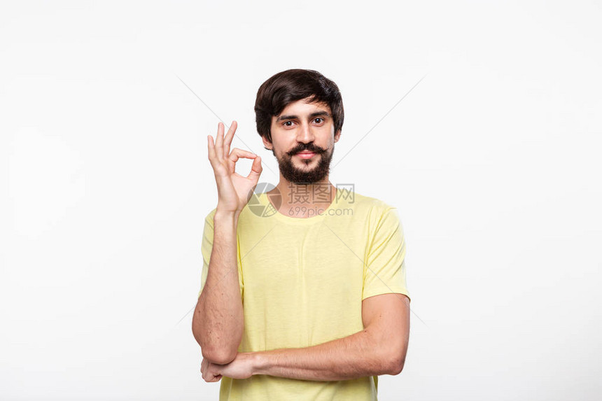 长着胡子的棕发大胡子男人穿着黄衬衫手势是OK牌站在白色背景图片