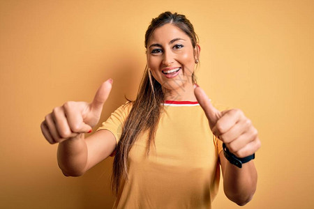 年轻漂亮的运动女穿着休闲t恤和马尾辫在黄色背景上批准用手做积极的手势图片