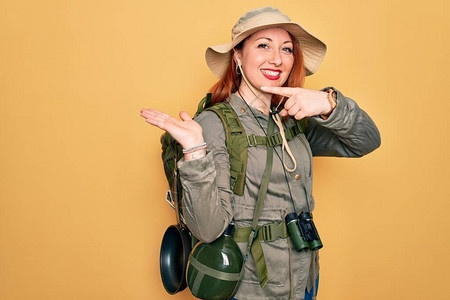 年轻的红发背包客女人穿着背包和帽子在黄色背景上徒步旅行图片