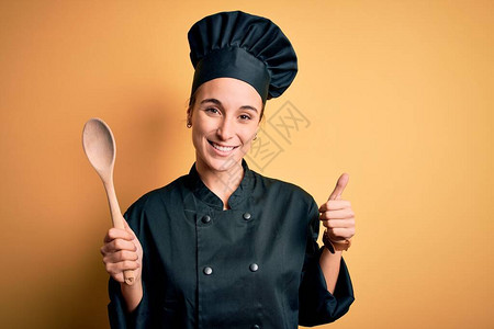 年轻漂亮的厨师女人穿着炊具制服和帽子拿着木勺开心地笑着做好手势图片