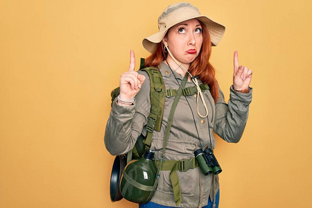 年轻的红发背包客女人在黄色背景下徒步旅行时背着包和帽子图片
