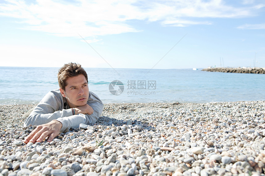 一个男人的肖像躺在白石子海滩的海岸上用蓝天空和海来深思熟图片
