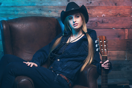 有原声吉他的女牛仔乡村歌手坐在皮椅上穿着蓝色牛仔裤和棕色图片