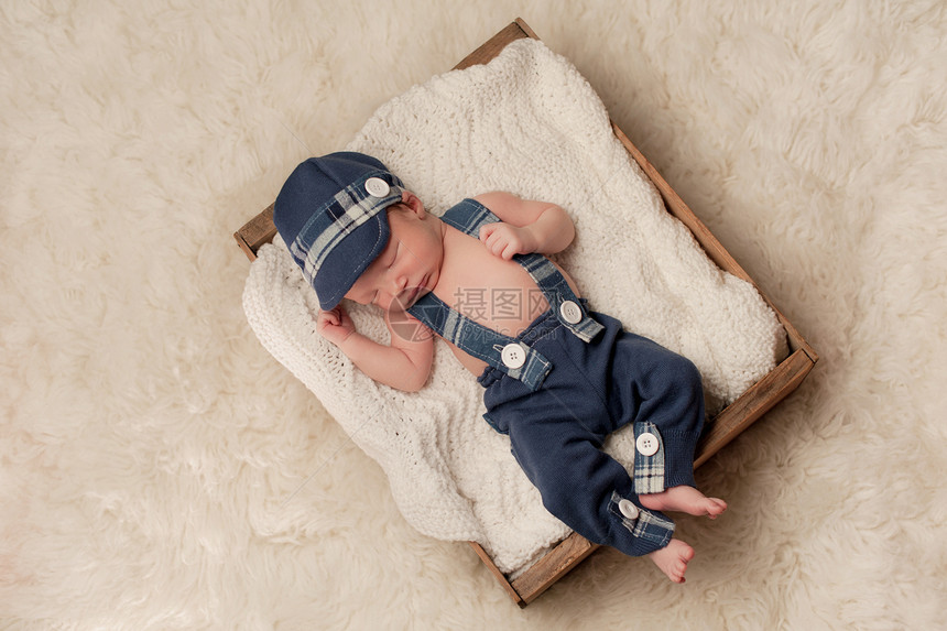 一个八天大的新生男婴戴着蓝色报童帽吊带和裤子的头顶照片他睡在一个旧木箱里在工作室的奶油色flokati图片