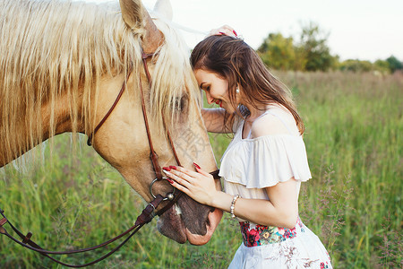 穿着白色裙子的迷人年轻女子站在绿草地上仰着美丽的白马和棕图片