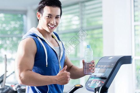 在运动场体育馆微笑的年轻男子在运动机上展示水瓶图片