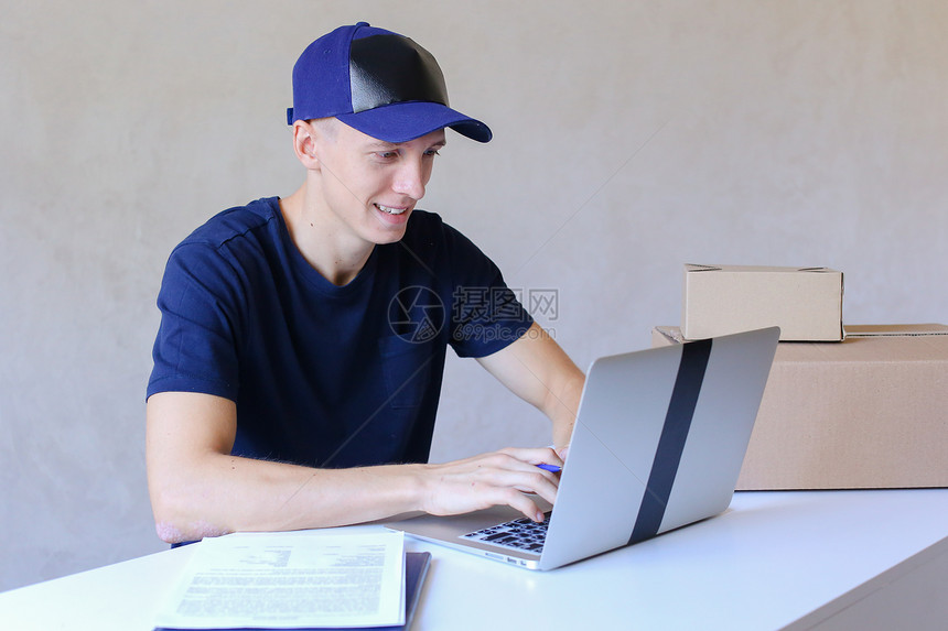 年轻的邮递员坐在桌前用笔记本电脑打字快递公司每天接受订单不同类型和尺寸的年轻送货员欧洲外观穿着黑色T恤和帽图片