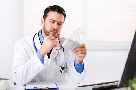 身穿白大衣颈部涂有听诊器的男医生坐在桌子上思考处方和阅读药剂标签图片