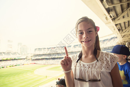 一个微笑的快乐小女孩在观看职业棒球比赛时显示出1号手指标志她正在享受在一个大球场观看她最喜欢背景