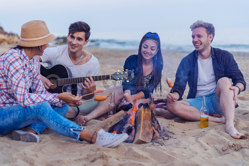 一群年轻开朗的朋友坐在沙滩上点燃篝火一名男子正在弹吉他野生海滩上的音乐图片