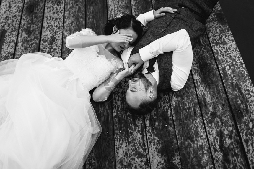 穿着优雅礼服的美丽奢华豪新娘和有胡子的新郎躺在雨中湖泊附近的一个木头码上从上面图片