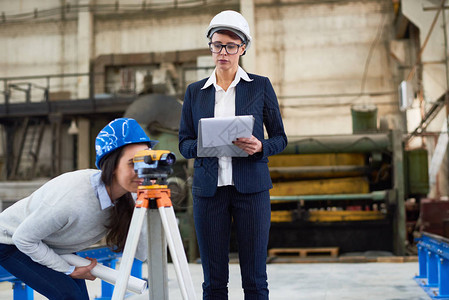 沉思的建筑测量师在与女工程师一起在工厂通过光学经纬仪观察时图片