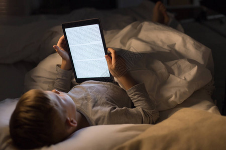晚上用电子阅读器在床上阅读书的小男孩高角度肖像图片