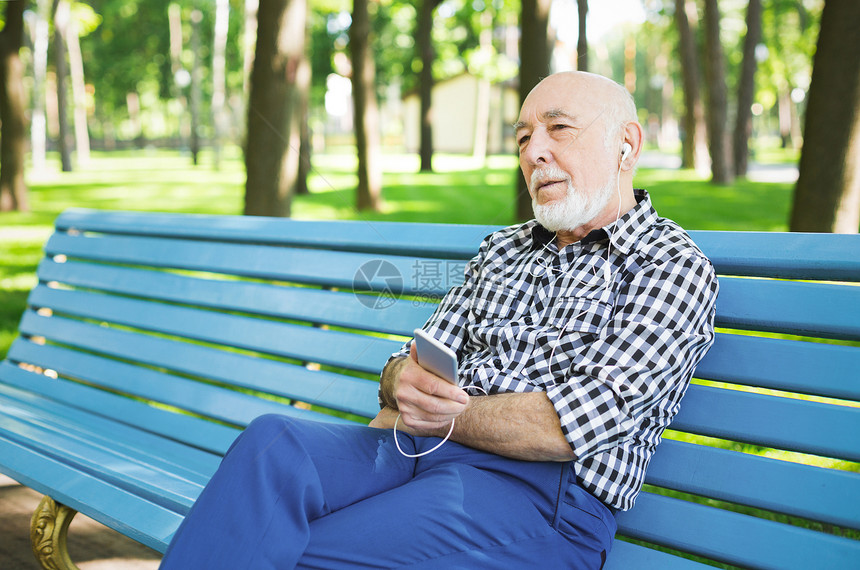 放松的老人在户外听音乐戴耳机的老人坐在公园的长椅上欣赏智能手机的音图片