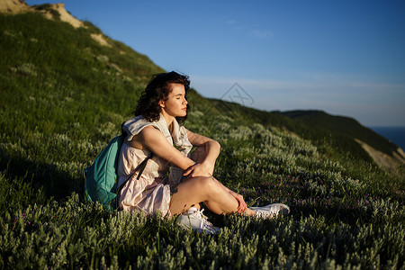 美丽的黑发女人坐在海山上的草地上带着背包在海洋附近旅行的女人旅行图片