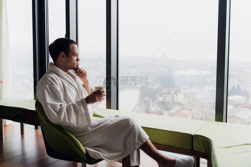 穿着浴袍的男人在靠近全景窗户的豪华摩天大楼酒店喝咖啡早上度假图片