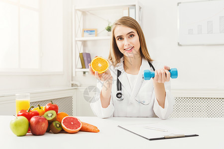 年轻的营养师在诊所用橙色和哑铃微笑的女营养师拿着水果和运动器材健康饮图片