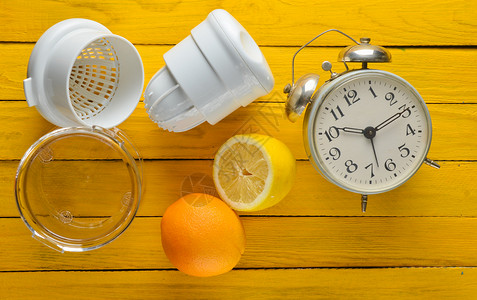 早晨从柠檬和橙子中提取的新鲜果汁手工榨汁机闹钟黄色木质背景上的柑橘类图片