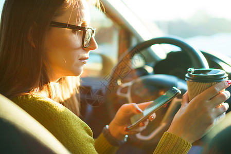 年轻妇女坐在车里时使用智能手机喝图片