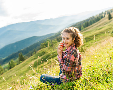 卷曲微笑的年轻女子在夏天坐在美丽的山景上与电话交谈现代人无论走到哪里都使用便背景图片