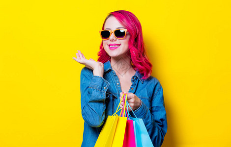 戴着太阳镜和蓝色衬衫的年轻粉红色头发女孩拿着彩色购物袋孤立的黄色图片