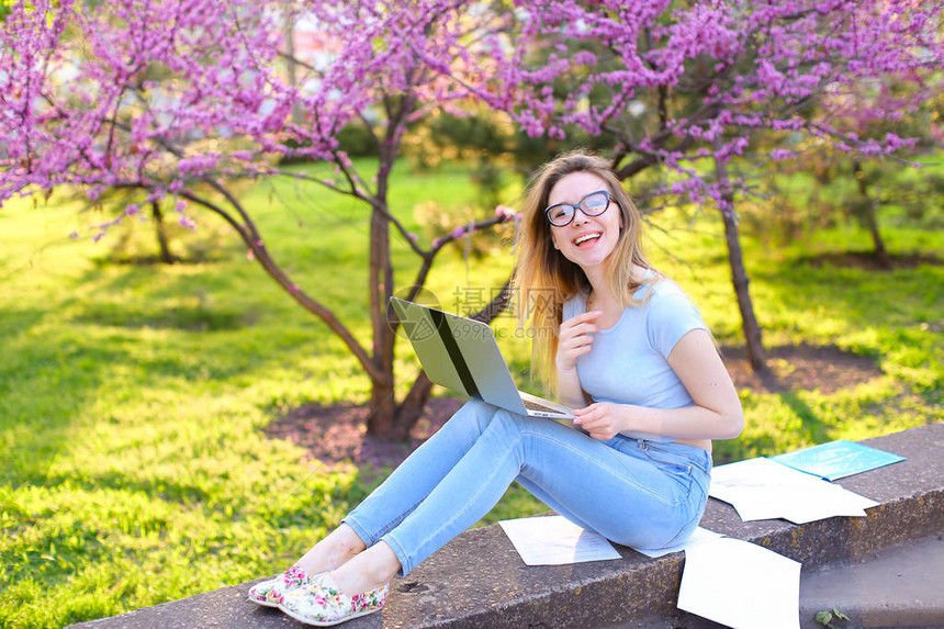 戴眼镜的快乐女学生准备在春季公园用笔记本电脑和论文进行考试图片