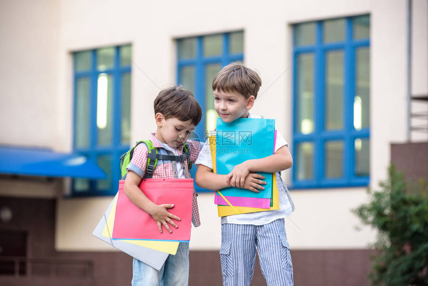 可爱的小学生在校园里轻快地交谈孩子们的心情很好温暖的春天早晨在同学的书包背后在其中一个男孩鲜红色图片